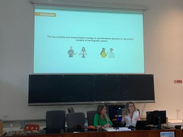 Two women sit at a table in front of a projection screen displaying a conclusion slide about iconicity in communication. The woman on the right, A. Slonimska, is speaking. There are snacks and water bottles on the table.