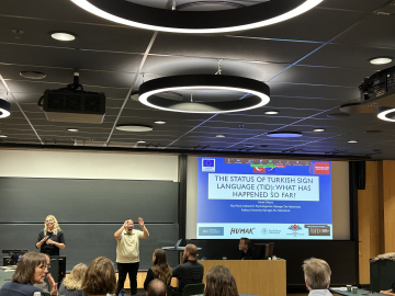 A conference room with a presentation on "The Status of Turkish Sign Language (TID): What Has Happened So Far?" Two presenters are on stage, one signing and the other speaking.