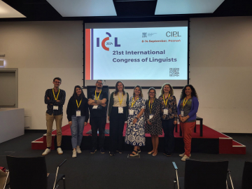 A group of eight participants at the 21st International Congress of Linguists poses for a photo on stage, all wearing conference badges and yellow lanyards, standing in front of the event's display screen.