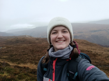 A woman smiling warmly, wearing a white hat and a dark gray jacket with an orange zipper, standing in front of a softly blurred mountainous background.