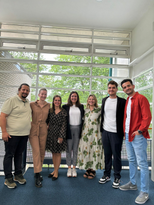 A group of seven people stand together, smiling, in front of a large window. They are dressed in a mix of casual and semi-formal attire. The background shows trees and a building exterior through the window.