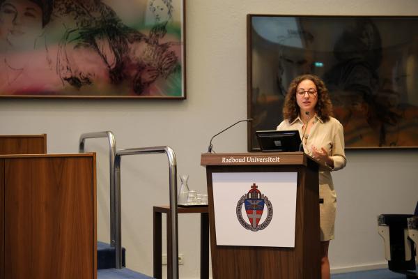 A woman with curly hair speaks at a podium marked with the Radboud Universiteit emblem in a conference room. Behind her, abstract paintings adorn the walls, enhancing the academic setting. She gestures with her hands, indicating an engaging discussion or presentation.   