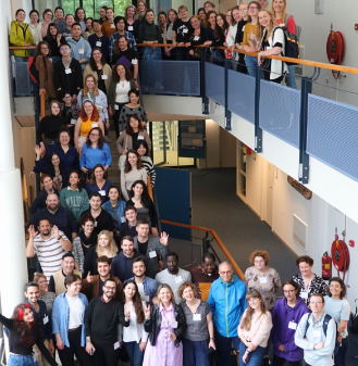 A large group of diverse individuals, gathered on a staircase and hallway of a building, posing for a group photo during the Medal summer school.