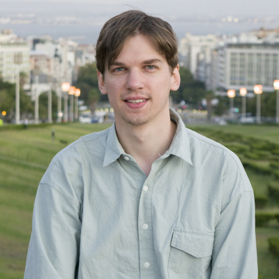 The image shows a young man with light brown hair, wearing a light green button-up shirt, standing outdoors with a cityscape and green parkland in the background.