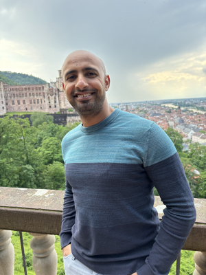 A man wearing a light and dark blue sweater stands smiling on a balcony with a view of city in the background.