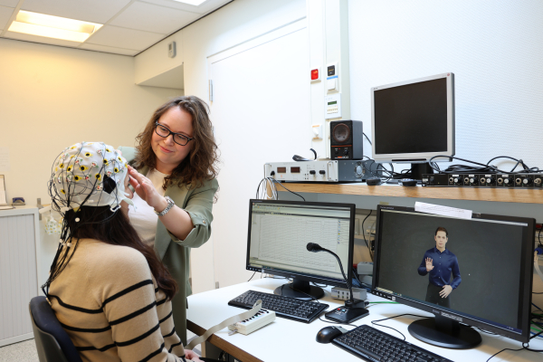 A researcher adjusts an EEG cap on a participant, surrounded by computers displaying EEG data and sign language software.