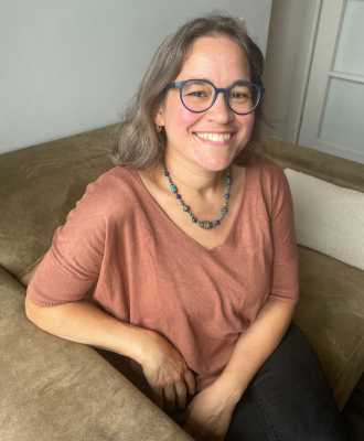 A smiling woman wearing glasses, sitting on a couch, in a cozy, casual setting indoors.