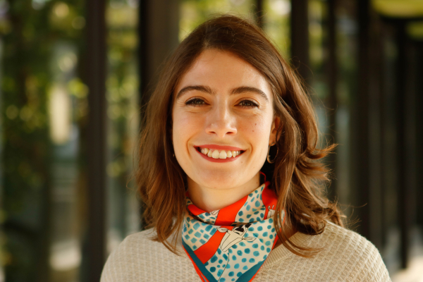  A woman smiling warmly, wearing a colorful scarf, standing in a softly blurred, green background.