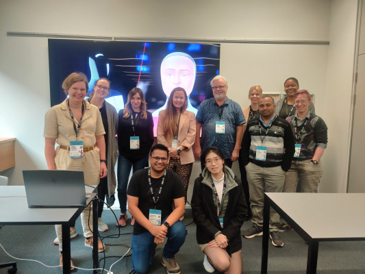  A group of eleven people pose together in a workshop room with a digital screen in the background.