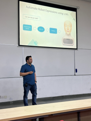  A man in a blue polo shirt and jeans presents in a lecture hall, standing near a whiteboard. Behind him, a slide titled "Automate Robot Expressions using LLMs" shows a flow diagram and a robotic face.